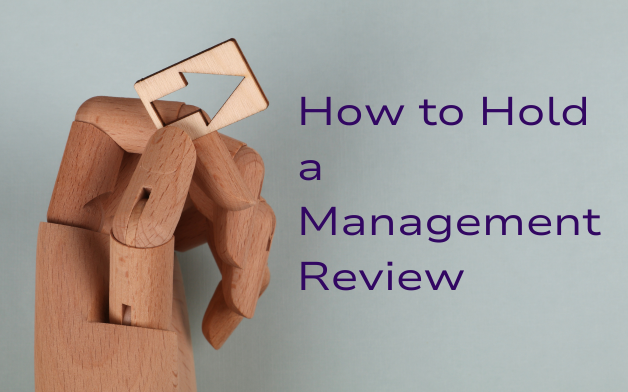 A wooden mannequin hand holding a square wooden block with an upward-pointing arrow cutout. The background is a soft gray-blue, and the text "How to Hold a Management Review" appears to the right in bold, purple font.